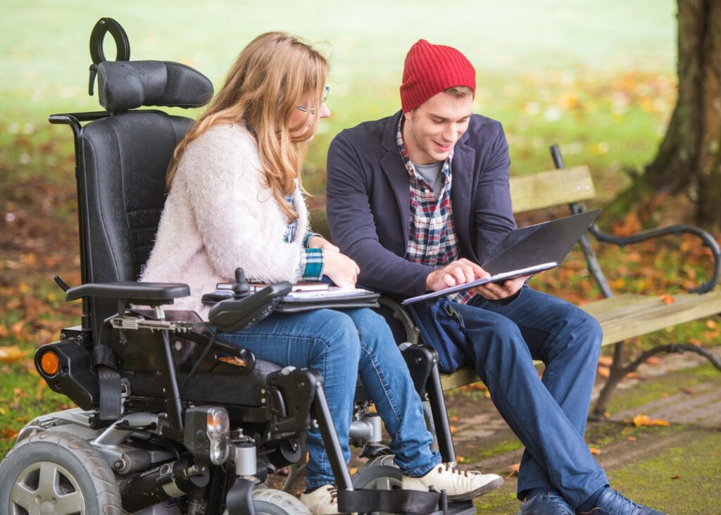 A white woman in a pink sweater and jeans is sitting in a wheelchair next to a white man in jeans, a gray sweater, a plaid shirt, and a red beanie hat. The man is sitting on a park bench